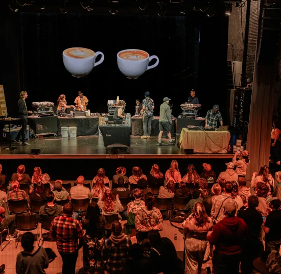 On a stage, baristas work on their latte art during a throwdown. A dj plays at one table, while 3 espresso machines are set up around the stage.