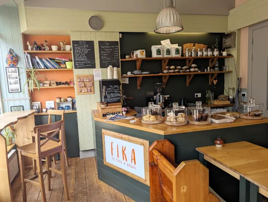 The bar, tables, stools, shelves and chairs inside Fika are of blonde wood, with muted tones of gray and green on the walls. Open shelves show books, coffee bags, teas, and pastries.