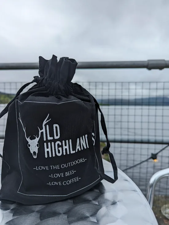 On a metal table outside facing the sea sits a black drawstring bag with a deer skull and antlers, logo that says Wild Highlands, and the words "love the outdoors, love bees, love coffee."