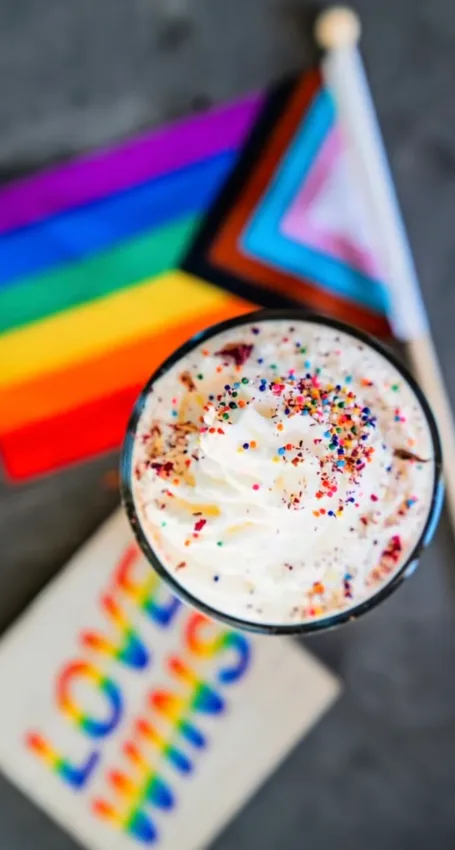 Top view of a latte with rainbow sprinkles on top and pride flag and love wins sign blurred in the background.