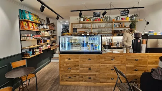 Inside Dutch Café. The bar is wood with small brass handles on its drawers. A glass case holds cold drinks. Open shelves hold coffee and food products. Plants hang from the ceiling.