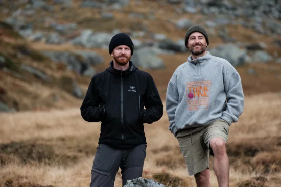 Two men with beards and knit hats stand in a rocky area.