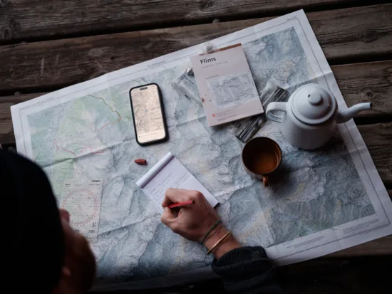 A person with coffee cup and a white pot looks over maps and takes notes in a small notebook.