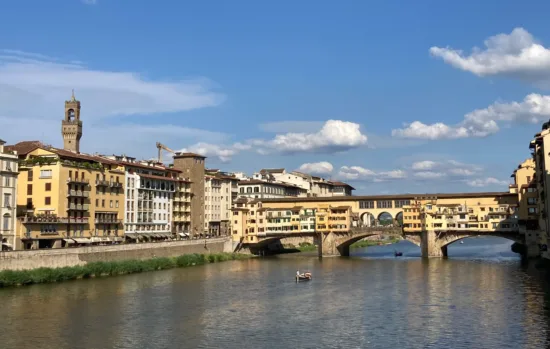 A view of the arched Ponte Vecchio bridge over the wide Arno, apartment buildings, and a slightly cloudy blue sky.