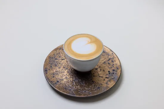 A small latte with heart shaped latte art, placed on a textured blue and gold saucer.