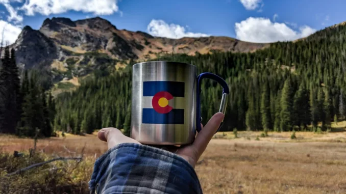 A person holds a coffee cup with the Colorado state flag on it in front of mountains.