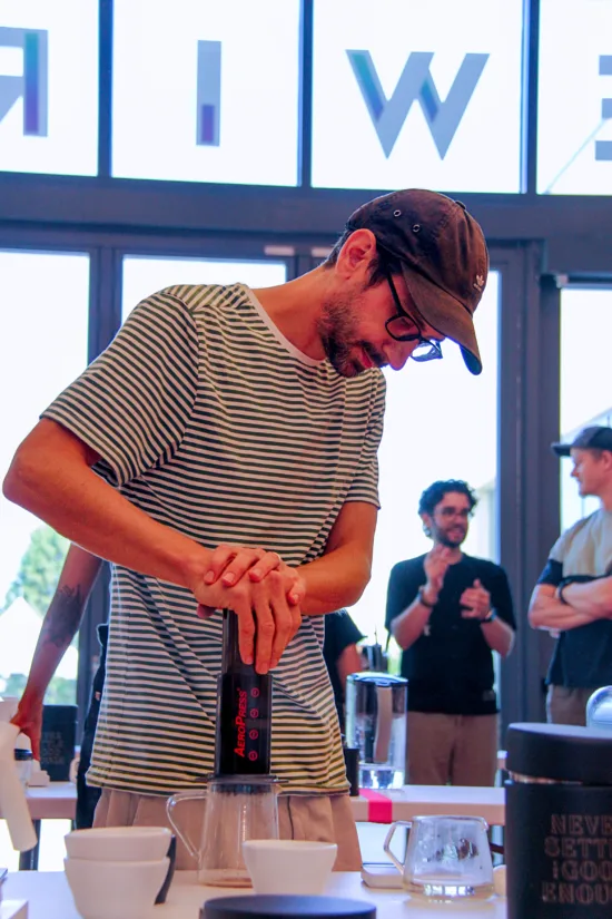 Dakota wears a baseball cap and striped shirt, and works the Aeropress.