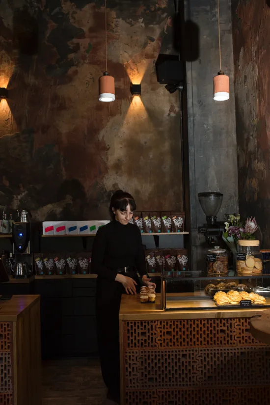 The dark brown interior of sensory Room, with a barista wearing black and holding pastries behind the decorative wooden counter.