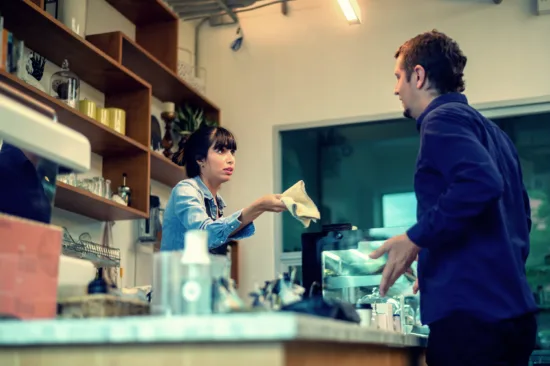 A barista stands behind a counter holding a bar towel, while a customer argues with her.