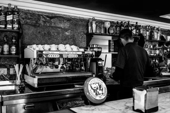 A barista works behind a counter tamping coffee next to an espresso machine and liquor shelf.