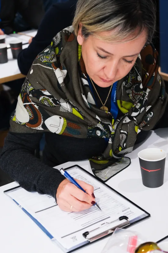 A person wears a shawl and writes on paper clipped to a clipboard. There is a black paper cup in front of them.