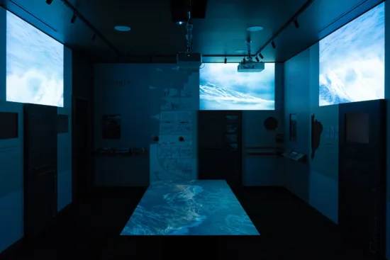 A view of the Coffee Migrant | Migrant Coffee exhibit, which shows a dark room with screens showing blue and white waves. Screens are on the walls and rising up from a table near the floor.