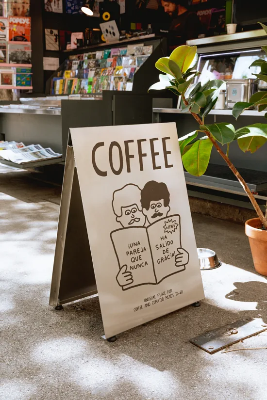 A sandwich saign says COFFEE and features two illustrated men with mustaches sharing a magazine. A fig tree is potted beside it with a newsstand behind.