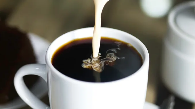 Milk being poured into a white mug of hot coffee.