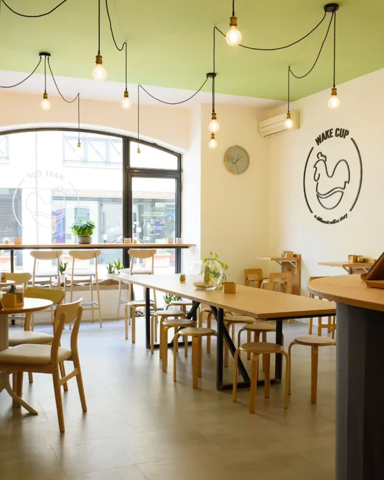 The sunlit interior of Wake Cup with light wooden furnishings, large window, and a spring green ceiling. A wake Cup sign with a rooster illustration is painted on the wall.