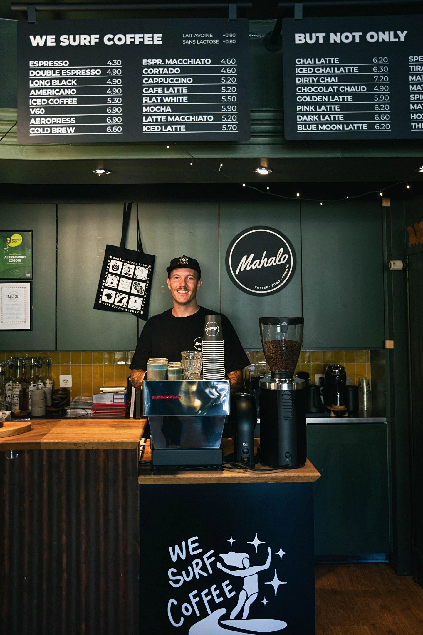 Jerome behind the bar at Mahalo's Fribourg location, which has black and green walls and counter. A sign featuring a surfer and the words "We Surf Coffee" is painted in front of the espresso machine.