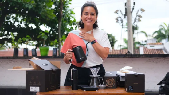 Maria holds a kettle and wears a GCAG wristband. On a table in front of her are other brewing implements: pourover brewers, scale, filters, and carafe with lid.