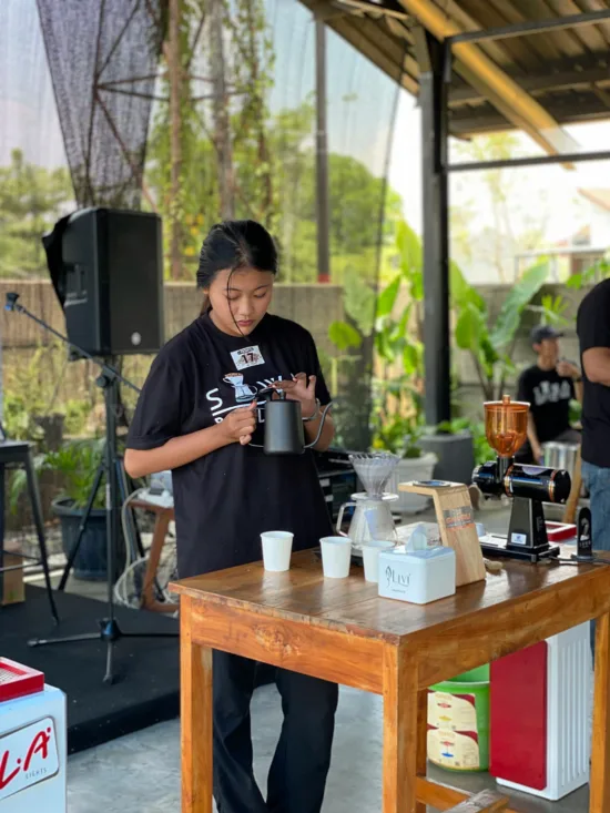 A barista holding a kettle with long spout. She is in an open-air shop.