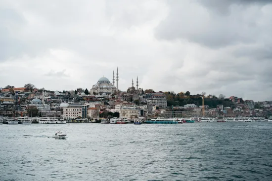 A boat on the waterfront of Istanbul. A large mosque and city landscape are seen in the nackground under an overcast sky.